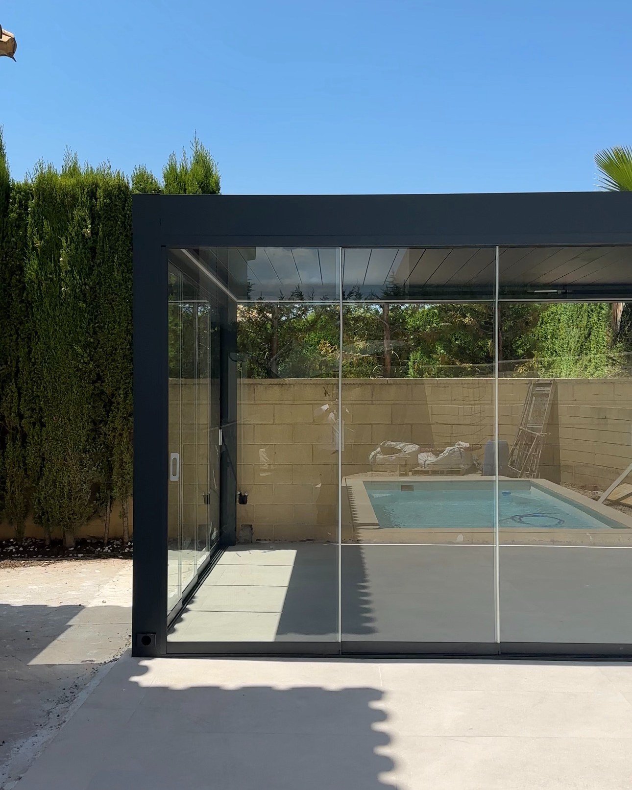 Vista frontal de la pérgola bioclimática con cortinas de cristal y la piscina al fondo